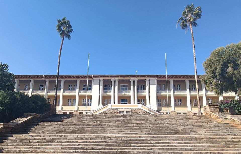 Der "Tintenpalast" / das Parlament in Windhoek, Namibia. Der Name kommt daher, dass in der Kolonialzeit viel Bürokratie herrschte und dort deswegen viel Tinte verschrieben wurde. Er zeigt, dass das aus Deutschland exportierte politische System den damaligen Bewohner*innen Namibias fremd war.